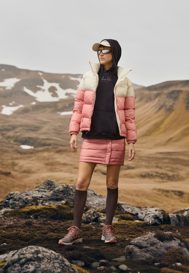 Puffermantel in Creme und Pink, über einem schwarzen Hoodie getragen. Pinke gestepfte Röcke, braune Kniestrümpfe und graue Sneaker mit pinken Akzenten.