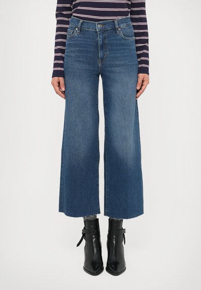 Jean large taille haute en denim bleu moyen, avec une texture délavée, un ourlet brut et des poches avant, assorti à des bottines noires.