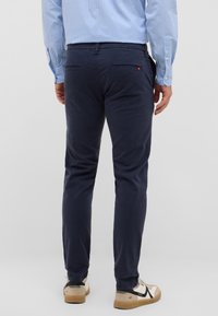 Marineblaue Baumwollhosen mit schlanker Passform, zwei Gesäßtaschen und einem kleinen roten Logodetail. Kombiniert mit beige und schwarzen Schuhen.