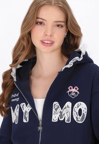 Sudadera con cremallera azul marino con adorno de encaje en la capucha, letras blancas bordadas "MY MO" y un pequeño diseño de corazón rosa. Textura suave.