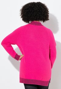 Persona con capelli ricci che indossa un maglione a maniche lunghe di un luminoso colore rosa con polsini e orlo a strisce nere, in piedi con le spalle rivolte verso di noi e una mano sulla vita.