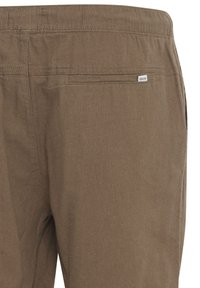 Brune stofbukser, der viser bagkanten og en enkelt ombukket lomme med en lille hvid etiket på højre side.