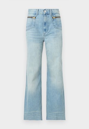 Γυναίκα με ανοιχτό μπλε τοπ, ανοιχτό μπλε τζιν με ψηλοκάβαλα και φαρδιά μπατζάκια με καφέ ζώνη, λευκές κάλτσες και καφέ μοκασίνια, στέκεται με το ένα χέρι στη μέση.