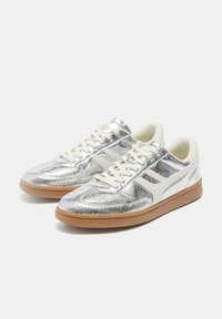 Pár alacsony szárú, fémszürke sneaker fehér fűzőkkel, krémszínű belsővel és gumitalpakkal, fehér háttéren.