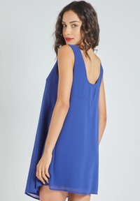 Mujer con cabello oscuro y ondulado que lleva un vestido azul real sin mangas con un escote en la espalda, mirando por encima del hombro contra un fondo sencillo.