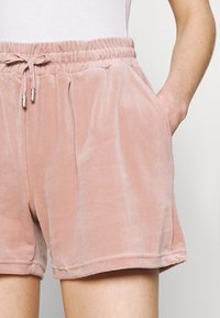 Short en velours rose doux avec une taille élastique, fermeture à cordon, poches latérales et coupe décontractée. Texture lisse et ourlet net.