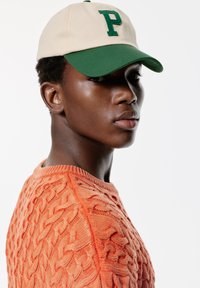 Cască bej cu margine verde și logo-ul brodat "P", asociată cu un pulover portocaliu cu tricotaj cu cablu, având modele texturale.