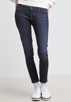 Vaqueros ajustados de mezclilla en color azul oscuro, con cierre de botón, bolsillos delanteros y detalles de costura sutiles. Combinados con zapatos y calcetines blancos.