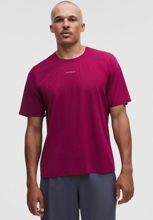 Mężczyzna z ogoloną głową, ubrany w gładki magentowy t-shirt z krótkim rękawem i ciemnoszare spodnie na jednolitym, jasnym tle.