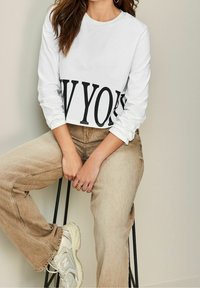 Frau, die auf einem Hocker sitzt, trägt ein weißes, cropped Sweatshirt mit dem Text "NEW YORK", beiges Jeans, weiße Sneakers und goldene Creolen.