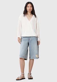 Hvid, struktureret wrap-bluse med lange ærmer, parret med lyseblå denim Bermuda-shorts med slidte detaljer. Sølvfarvede sandaler fuldender looket.