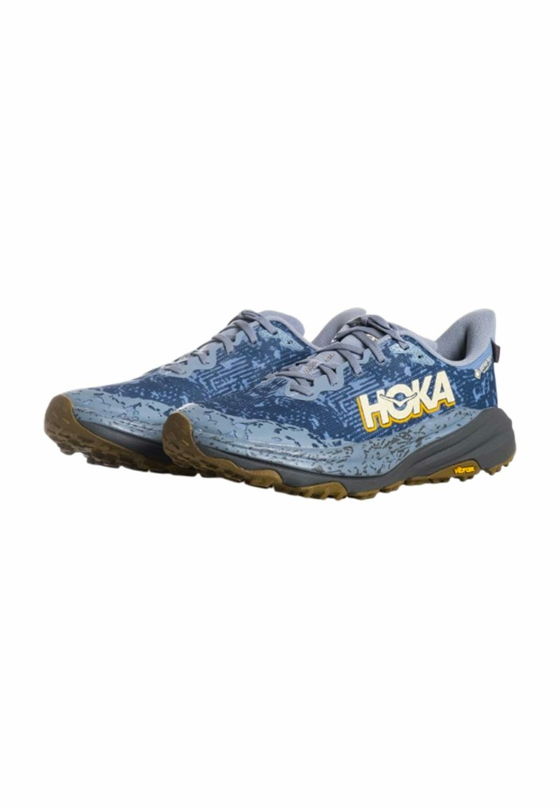 Coppia di scarpe da trail running HOKA blu con lacci grigi, tomaia con motivo e suole Vibram marroni, inclinate verso destra su sfondo bianco.