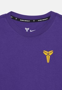 Violetinio medvilninio T-shirt su geltonu logotipu krūtinėje ir juodu kaklu, kuriame yra tekstas "MAMBA MENTALITY". Lygus paviršius.