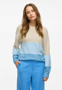 Lekki sweter dziewiarski w beżowe i niebieskie poziome pasy, z luźnym krojem i ściągaczami. W zestawie z niebieskimi spodniami.