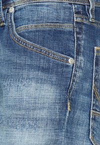 Vaqueros de mezclilla azul con desvanecimiento ligero, textura visible, costuras amarillas en contraste, detalle del bolsillo delantero y acento de botón metálico.