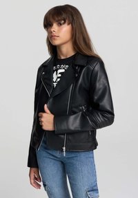 Giovane con lunghi capelli castani indossa un giubbotto di pelle nera sopra una maglietta con grafica e jeans cargo blu, tenendo il bavero della giacca con la mano sinistra.