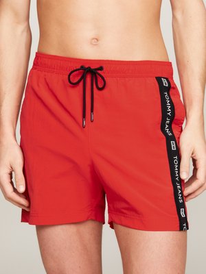 Bañador rojo con cinturilla elástica y cordón negro. En el lateral presenta la marca 'TOMMY JEANS' en blanco sobre una franja negra. Tela ligera.