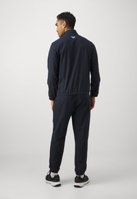 Marineblauw trainingspak in een gestructureerde stof, met een ritsjack en elastische boorden, gecombineerd met bijpassende joggers en sportschoenen.