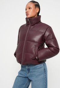 Veste en simili cuir matelassée bordeaux avec un col montant, fermeture éclair à l'avant et poches latérales, associée à un jean en denim bleu clair.
