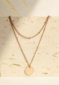 Różowo-złoty naszyjnik z dwiema warstwami łańcuszków i okrągłym zawieszką. Zawieszka jest gładka i nieozdobiona, ustawiona na tle teksturowanego kamienia.