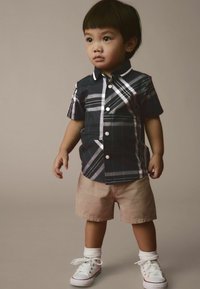 Camicia a maniche corte a quadri blu navy con accenti bianchi, abbinata a pantaloni corti beige e sneaker bianche, con manicotti arrotolati e chiusura a bottone.