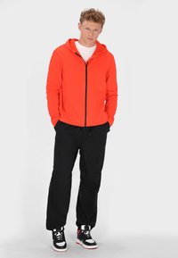 Světle oranžová mikina na zip s černým zipem, v kombinaci se volnými černými kalhotami a černo-bílými teniskami. Jednoduchý design, pohodlný střih.
