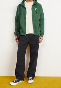 Grön zip-up hoodie med framfickor och en logo, bärs över en beige skjorta. Matchad med svarta vida byxor och svarta sneakers mot en gul bakgrund.