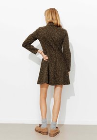 Robe courte vert olive à motif léopard, manches longues et taille cintrée, avec une jupe évasée. Portée avec des chaussures beige et des chaussettes grises.