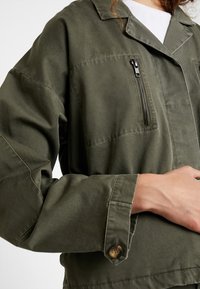 Oliivinvihreä takki, jossa on kaulus, vetoketjullinen rintatasku, napilla suljettavat hihansuut ja rento malli. Valmistettu kestävästä kankaasta.