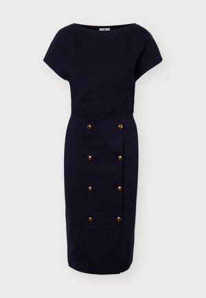 Femme portant un haut bleu marine à manches courtes et une jupe assortie longueur genou avec des boutons dorés, debout devant un fond clair uni.