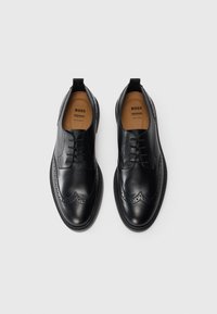 Sorte lærdressko med brogue-detaljer, med en glat finish, snørebåndsdesign og en rund tåform. Lysebrune indersåler synlige.