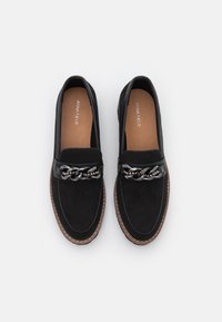 Mocassins noirs avec une tige en daim, une chaîne brillante en accent sur le devant, et une semelle intérieure en cuir tan avec des détails cousus.