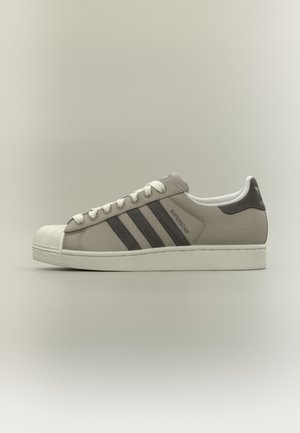 Chaussure en maille grise avec embout en caoutchouc blanc, trois bandes gris foncé et lacets blancs. Présente une tige texturée et un marquage signature.