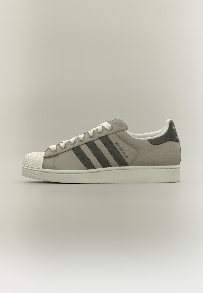 Chaussure en maille grise avec embout en caoutchouc blanc, trois bandes gris foncé et lacets blancs. Présente une tige texturée et un marquage signature.