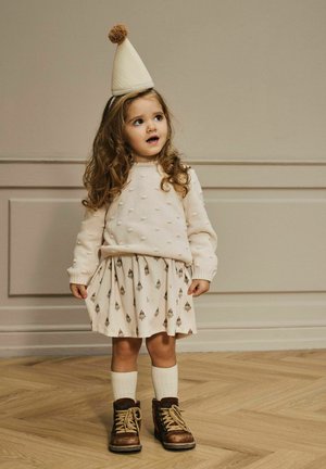 Junges Mädchen mit langen lockigen Haaren, das einen cremefarbigen Pullover, einen gemusterten Rock, kniehohe Socken, braune Stiefel und eine Partykappe mit einem Pompon trägt.