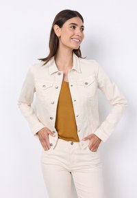 Giacca di denim beige con bottoni argentati, due tasche sul petto e maniche lunghe, indossata sopra una maglietta a V color senape e pantaloni beige chiaro.