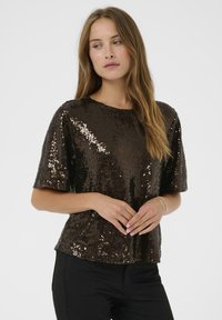 Vrouw met lang lichtbruin haar, gekleed in een zwarte korte mouwen top met sequins en zwarte broek, kijkend opzij met handen gevouwen.