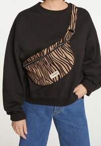 Personne portant un jean bleu et un sweatshirt noir avec un sac bandoulière zébré beige et noir, muni d'une fermeture éclair et d'une étiquette.
