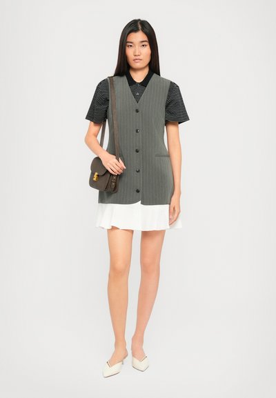 Femme portant un polo à rayures noires sous une robe débardeur à rayures gris avec un ourlet en volants blancs, portant un sac à bandoulière marron.