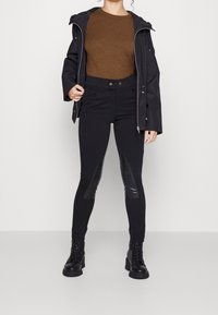 Frau trägt einen braunen Pullover, eine schwarze Reißverschlussjacke, schwarze Hosen mit Lederknieauflagen und schwarze Schnürstiefeletten, steht an einer weißen Wand.