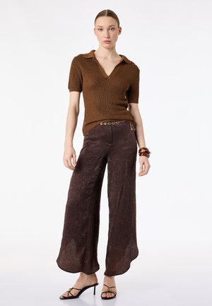 Femme debout portant un haut marron en maille à manches courtes, un pantalon ample texturé, des sandales noires à talons, une ceinture chaîne dorée et des bracelets au poignet droit.