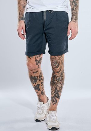 Mann trägt hochgekrempelte schwarze Shorts, weiße Sneaker und ein weißes T-Shirt und zeigt detaillierte Tätowierungen an beiden Beinen und Armen von den Knien bis zu den Handgelenken.