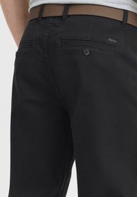 Pantalons en coton noirs avec une texture lisse, deux poches arrière avec boutons, et une étiquette logo discrète sur la poche droite.