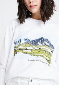 Kobieta w białej bluzie z napisem "WSPIERAJ SWOJĄ LOKALNĄ PLANETĘ" oraz krajobrazem górskim.