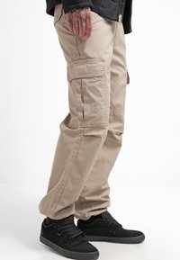 Beige cargo pants made of cotton, featuring side pockets and a relaxed fit. Worn with black sneakers, highlighting textured fabric.
