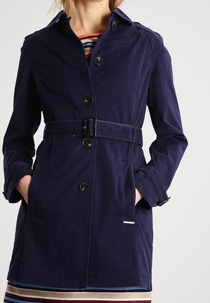 Kobieta nosząca granatowy trencz z paskiem i czarnymi guzikami, ręce w kieszeniach, na kolorowej, pasiastiej sukience.