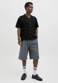Chemise noire à manches courtes avec un motif texturé, shorts gris, chaussures noires et chaussettes blanches avec des rayures noires, se tenant devant un fond neutre.