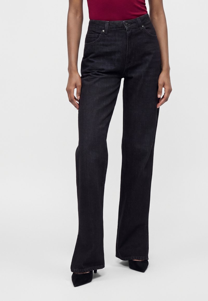 Personne portant un pantalon en denim noir taille haute à jambes droites et des talons noirs à bouts pointus, associé à un haut rouge.