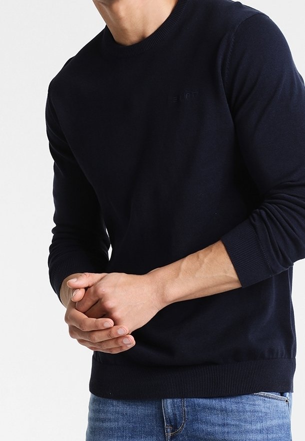 Homme portant un pull bleu marine à manches longues et un jean bleu, les mains lâchement jointes devant un fond clair uni.