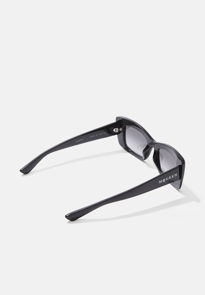 Crne plastične naočale za sunce s odvažnim, kutnim oblikom. Tamne leće i elegantne naramnice sadrže diskretno brendiranje s jedne strane.